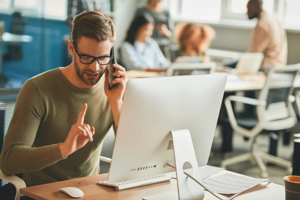 Home 9 Fotograf Hannover - Ein Mann mit Brille sitzt an einem Schreibtisch in einem Büro, telefoniert mit seinem Smartphone und gestikuliert mit dem Zeigefinger. Sein Blick ist auf einen Computermonitor gerichtet, umgeben von Papieren und einer Tastatur. Während andere in der Nähe arbeiten, überlegt er, ob er für das kommende Projekt einen „Fotografen in der Nähe“ anrufen soll. Fotograf in der Nähe.