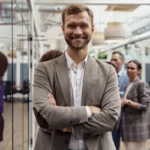 360 Grad Rundgang für Hotels 25 Fotograf Hannover - Ein Mann mit Bart und Lächeln steht selbstbewusst mit verschränkten Armen da und trägt einen beigen Blazer über einem weißen Hemd. In einem Bereich mit Glaspaneelen scheint er 360-Grad-Rundgänge zu planen, während sich im Hintergrund drei Personen unterhalten, die teilweise unscharf sind. Fotograf in der Nähe.