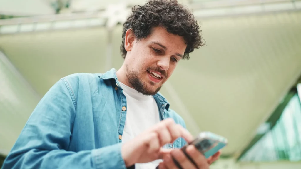 Was ist Google Street View? 22 Fotograf Hannover - Ein Mann mit lockigem Haar erkundet Google Street View auf seinem Telefon. Er trägt ein blaues Hemd über einem weißen T-Shirt. Hinter ihm ragt eine beeindruckende moderne Architekturstruktur mit ihren markanten Glas- und Metallelementen hervor. Fotograf in der Nähe.