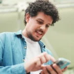 Was ist Google Street View? 7 Fotograf Hannover - Ein Mann mit lockigem Haar erkundet Google Street View auf seinem Telefon. Er trägt ein blaues Hemd über einem weißen T-Shirt. Hinter ihm ragt eine beeindruckende moderne Architekturstruktur mit ihren markanten Glas- und Metallelementen hervor. Fotograf in der Nähe.