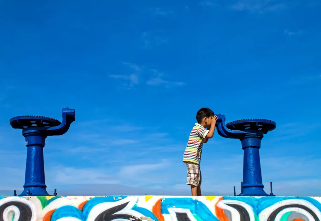 Wie funktioniert Google Street View? 20 Fotograf Hannover - Ein Kind in einem gestreiften Hemd blickt durch ein blaues Teleskop, das an einer mit Graffiti bedeckten Wand befestigt ist. Es erinnert an Szenen, die mit Google Street View aufgenommen wurden. Ein weiteres Teleskop steht ganz in der Nähe unter dem klaren, blauen Himmel. Fotograf in der Nähe.