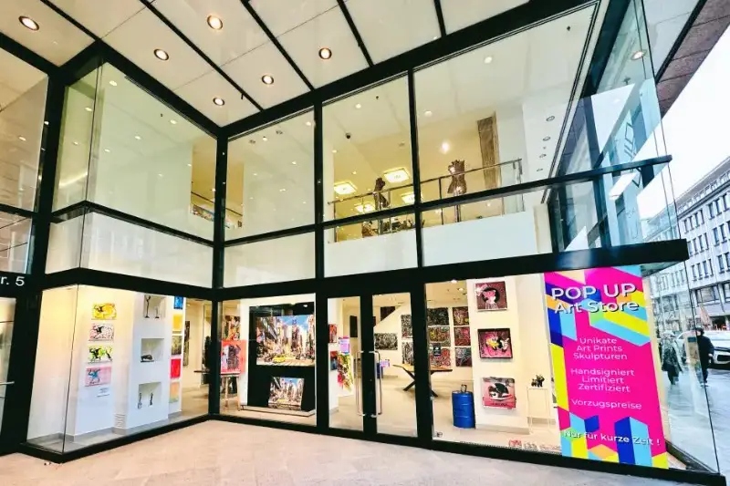 Produktfotos 15 Fotograf Hannover - Pop-up-Kunstladen mit Glasfront und farbenfrohem Schild, der einzigartige Kunstdrucke und Skulpturen anbietet. Im Inneren werden verschiedene Kunstwerke in einem gut beleuchteten Raum ausgestellt, der durch große Fenster sichtbar ist. Straße und benachbarte Gebäude spiegeln sich im Glas. Fotograf in der Nähe.