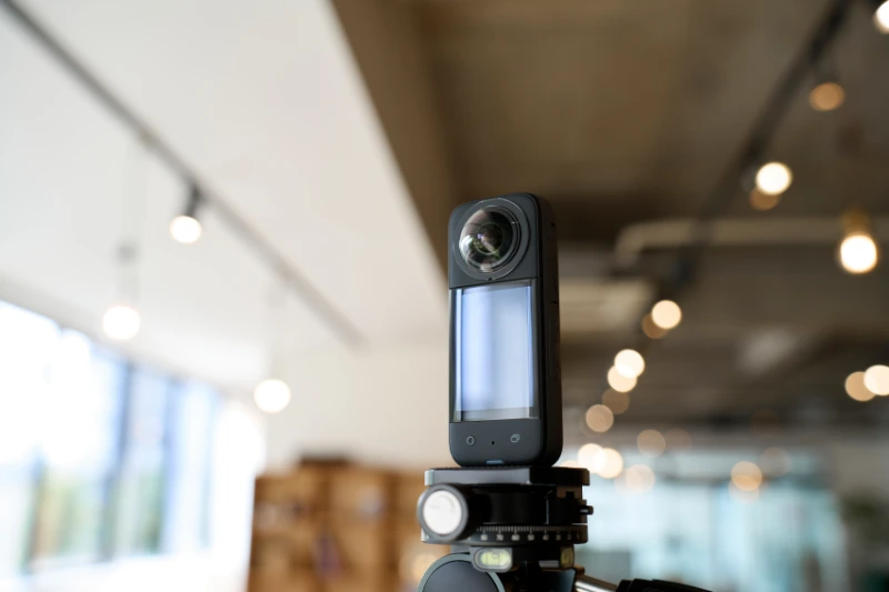 360 Grad Kamera Insta360 X3 Test 1 Fotograf Hannover - Eine schwarze Insta360 X3-Kamera auf einem Stativ ist im Innenbereich positioniert. Der Hintergrund zeigt einen modernen Raum mit freiliegenden Betondecken und Hängelampen, die kunstvoll verschwommen sind, um eine Tiefenschärfe zu erzielen. Das Objektiv und der kleine Bildschirm der Kamera fallen in diesem fokussierten Testaufbau auf. Fotograf in der Nähe.