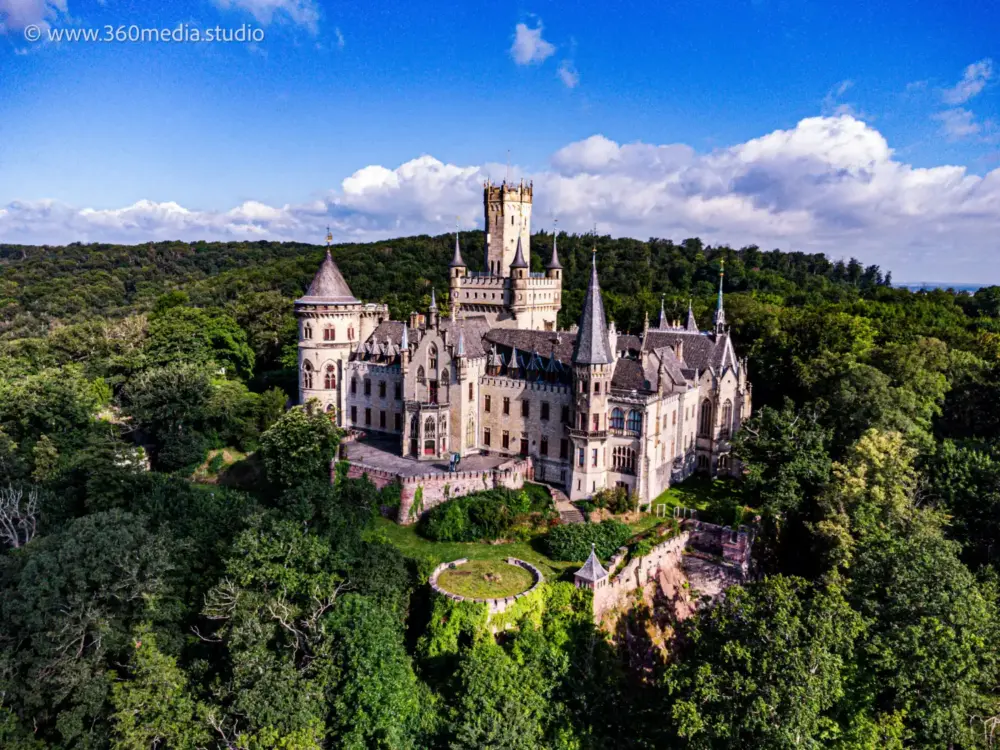 Produktfotos 14 Fotograf Hannover - Eine Luftaufnahme zeigt das majestätische Schloss Marienburg, ein historisches Juwel, umgeben von dichtem grünem Wald. Seine Türme haben spitze Dächer und komplizierte architektonische Details, und das alles unter einem hellen Himmel mit vereinzelten Wolken. Fotograf in der Nähe.