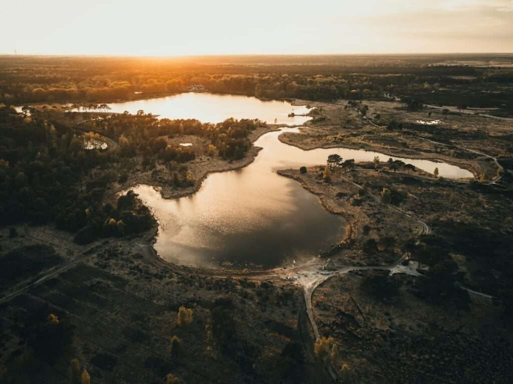 Drohnenaufnahmen: Die Vorteile der Luftbildfotografie 16 Fotograf Hannover - Luftaufnahme einer Landschaft bei Sonnenuntergang mit einem gewundenen Gewässer, umgeben von Bäumen und offenem Land. Die Sonne steht tief am Horizont und wirft einen warmen Schein auf die Szene. An den Rändern des Wassers ist spärliche Vegetation sichtbar. Fotograf in der Nähe.