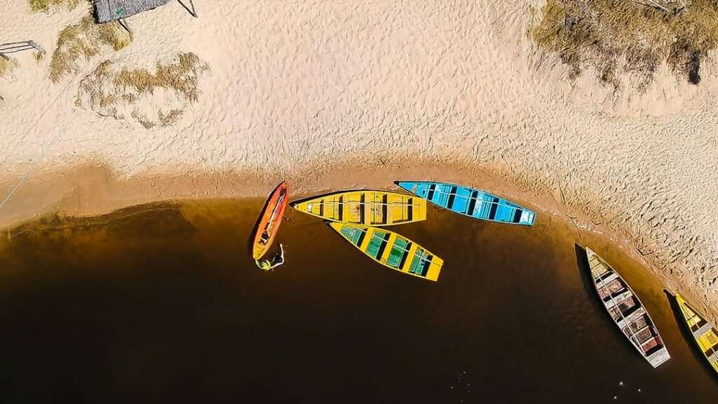 Die Kosten für Drohnenaufnahmen: Preise und Kalkulation 13 Fotograf Hannover - Luftaufnahme eines Sandstrands mit mehreren bunten Kanus, die am Wasserrand aufgereiht sind. Die Kanus sind rot, gelb und blau. Auf dem Sand ist etwas Gras zu sehen und die Uferlinie schlängelt sich sanft um die Boote. Fotograf in der Nähe.