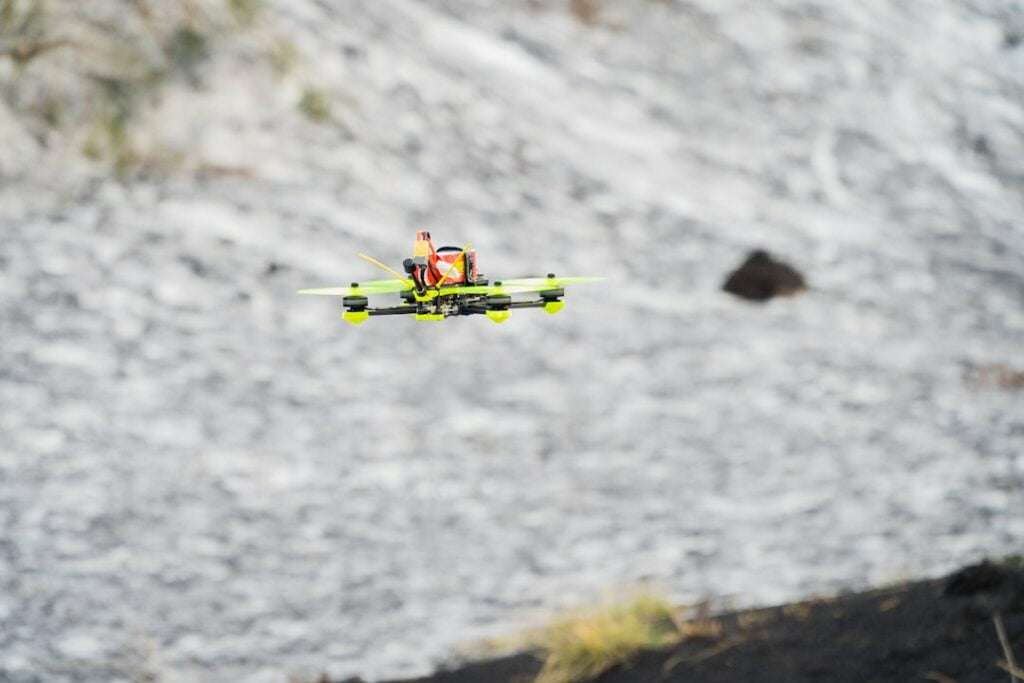 Sind Aufnahmen mit Drohnen erlaubt? 1 Fotograf Hannover - Eine kleine Drohne mit gelben Propellern, ausgestattet mit einer Kamera, fliegt über ein felsiges Gelände. Der Hintergrund ist etwas verschwommen, was auf Bewegung hindeutet. Die Landschaft wirkt karg, mit sichtbaren Grasflecken. Fotograf in der Nähe.