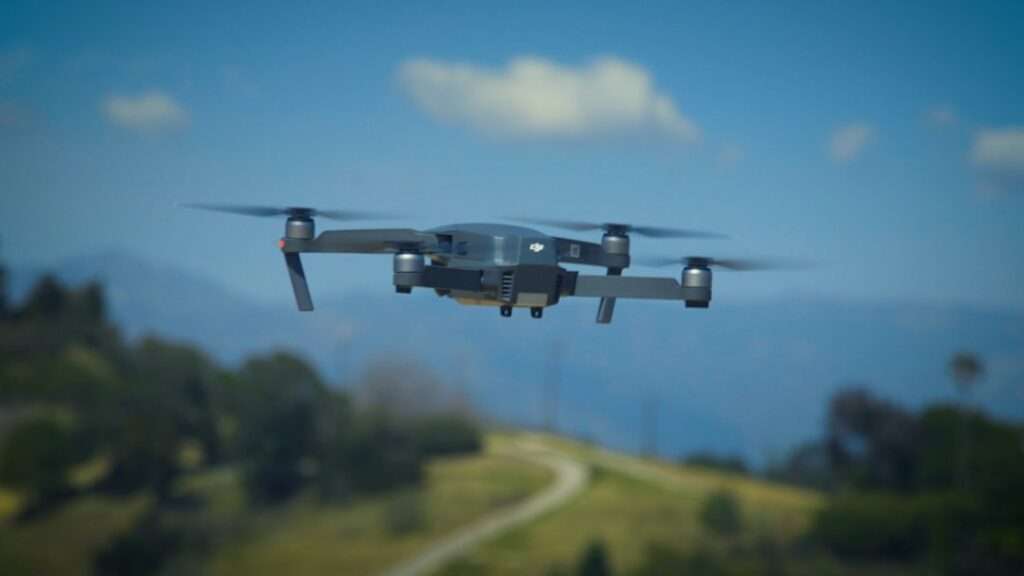 Kosten für Drohnenaufnahmen: Was Sie wissen müssen 10 Fotograf Hannover - Eine Drohne mit vier Propellern schwebt vor dem Hintergrund einer verschwommenen Landschaft in der Luft. Die Szene umfasst Hügel, einen gewundenen Pfad und einen blauen Himmel mit vereinzelten Wolken. Fotograf in der Nähe.