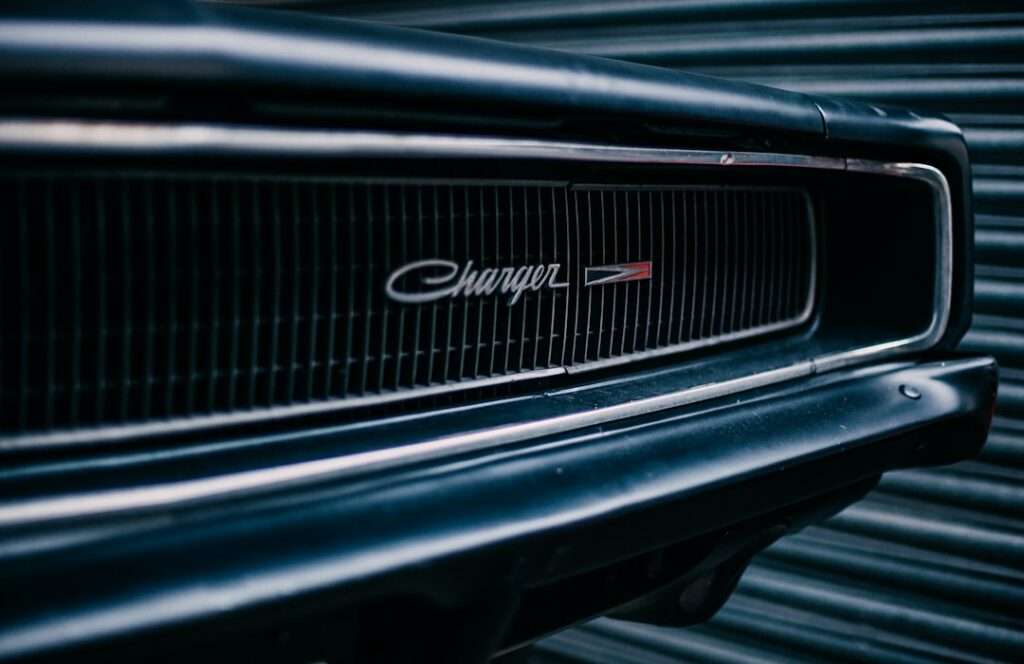 Autos fotografieren: Tipps und Tricks für tolle Bilder 17 Fotograf Hannover Nahaufnahme des Kühlergrills eines schwarzen Oldtimers. Er zeigt das „Charger“-Emblem mit einem rot-weißen Logo daneben. Der Hintergrund zeigt eine gewellte Metalloberfläche. Fotograf in der Nähe.