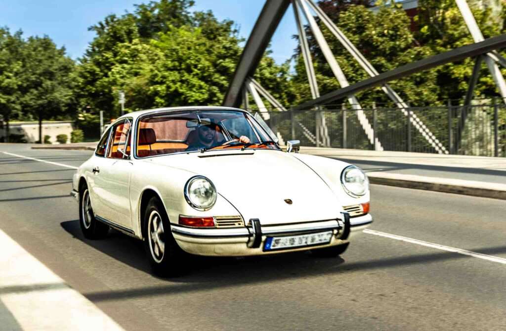Dein ultimatives Fotoshooting mit dem Auto 1 Fotograf Hannover Ein weißer Porsche-Oldtimer-Sportwagen mit runden Scheinwerfern fährt eine Stadtstraße entlang, im Hintergrund eine moderne Brücke und Bäume. Das Auto wurde von Auto Fotograf Hannover perfekt aufgenommen und hat ein blau-weißes europäisches Nummernschild. Der Fahrer ist im Auto zu sehen. Fotograf in der Nähe.