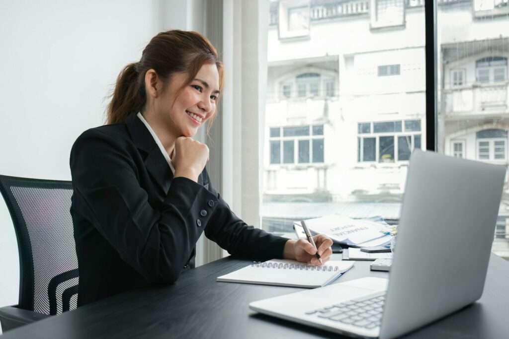 Was ist Corporate Photography? 4 Fotograf Hannover Eine Frau in einem schwarzen Blazer sitzt an einem Schreibtisch vor einem Laptop, lächelt und schreibt in ein Notizbuch. Auf dem Schreibtisch liegen Dokumente, im Hintergrund sieht man große Fenster mit Blick auf die Stadtlandschaft – die Essenz der Unternehmensfotografie. Fotograf in der Nähe.