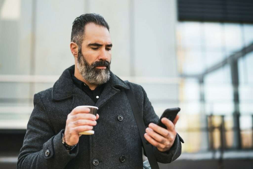 Professionelles Business Portrait Fotografie: Ein Muss für Erfolgreiche Unternehmer 12 Fotograf Hannover Draußen steht ein bärtiger Mann in einem dunklen Mantel. In der linken Hand hält er einen Pappbecher, in der rechten schaut er auf ein Smartphone. Das Porträt zeigt ihn vor dem Hintergrund von Glasfenstern und einem Teil der Außenfassade des Gebäudes. Fotograf in der Nähe.