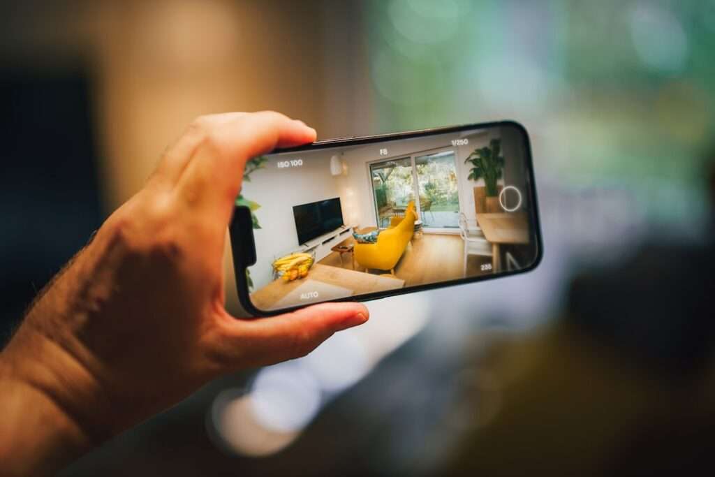 Entdecke die Stadt mit 360 Rundgängen 6 Fotograf Hannover Eine Hand hält ein Smartphone horizontal und macht damit ein Foto von einem modernen Wohnzimmer mit gelbem Sofa, Fernseher und großen Fenstern, die den Blick ins Grüne draußen freigeben. Der Hintergrund erscheint verschwommen – perfekt für die Erstellung immersiver 360-Grad-Rundgänge. Fotograf in der Nähe.