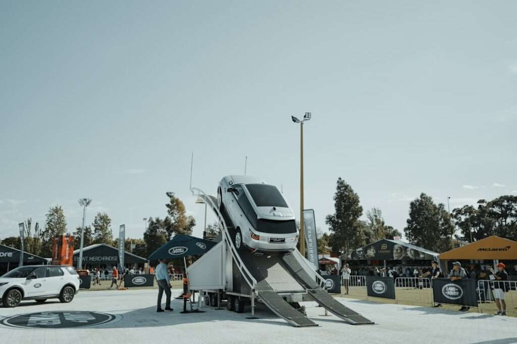 Fahrzeug Fotografie: Wie du deine Autoaufnahmen verbessern kannst 4 Fotograf Hannover Ein weißer SUV steht bei einer Outdoor-Automobilveranstaltung vertikal auf einer Rampe und präsentiert beeindruckende Fahrzeugfotografie. Eine Person steht in der Nähe, während im Hintergrund mehrere Stände von Automarken unter Zelten zu sehen sind. Bäume und ein klarer Himmel runden die lebendige Szene ab. Fotograf in der Nähe.