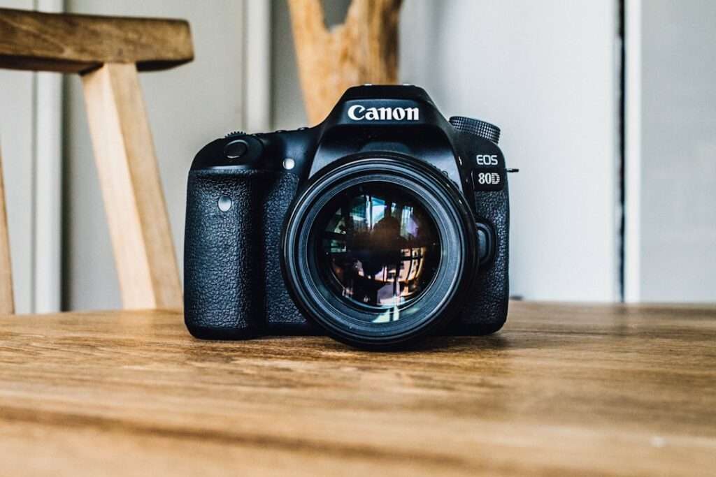 Entdecke die Welt mit Google 360 4 Fotograf Hannover Eine Canon EOS 80D DSLR-Kamera, in deren Linse eine detaillierte Spiegelung sichtbar ist, ruht auf einer Holzoberfläche. Im Hintergrund sind Teile eines Holzstuhls und eine weiße Wand zu sehen. Fotograf in der Nähe.