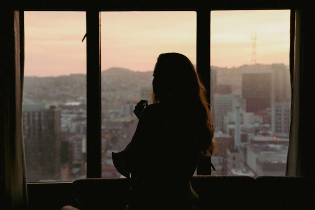Tipps für gelungene Hotel Fotografie 1 Fotograf Hannover Eine Person mit langen Haaren ist als Silhouette vor einem großen Fenster in ihrem Hotelzimmer zu sehen und blickt bei Sonnenuntergang auf eine Stadtlandschaft. Der Raum ist schwach beleuchtet und die Außenszene zeigt Gebäude und entfernte Hügel unter einem Himmel mit Farbverlauf, was einen Moment perfekter Fotografie einfängt. Fotograf in der Nähe.