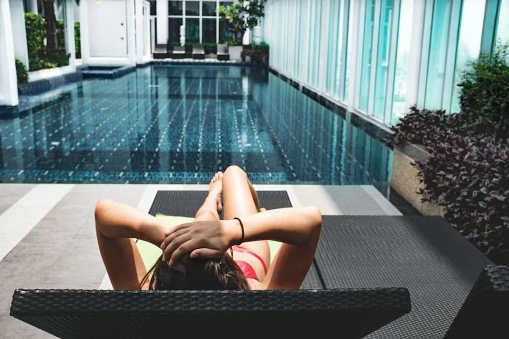 Entdecke das perfekte Hotel für Fotoshooting! 16 Fotograf Hannover Eine Person liegt mit verschränkten Händen auf einem Liegestuhl am Pool und blickt auf einen modernen Pool, der von Glaswänden und Grün umgeben ist. Die Person trägt Badebekleidung, wirkt entspannt und genießt die sonnige Umgebung. Fotograf in der Nähe.