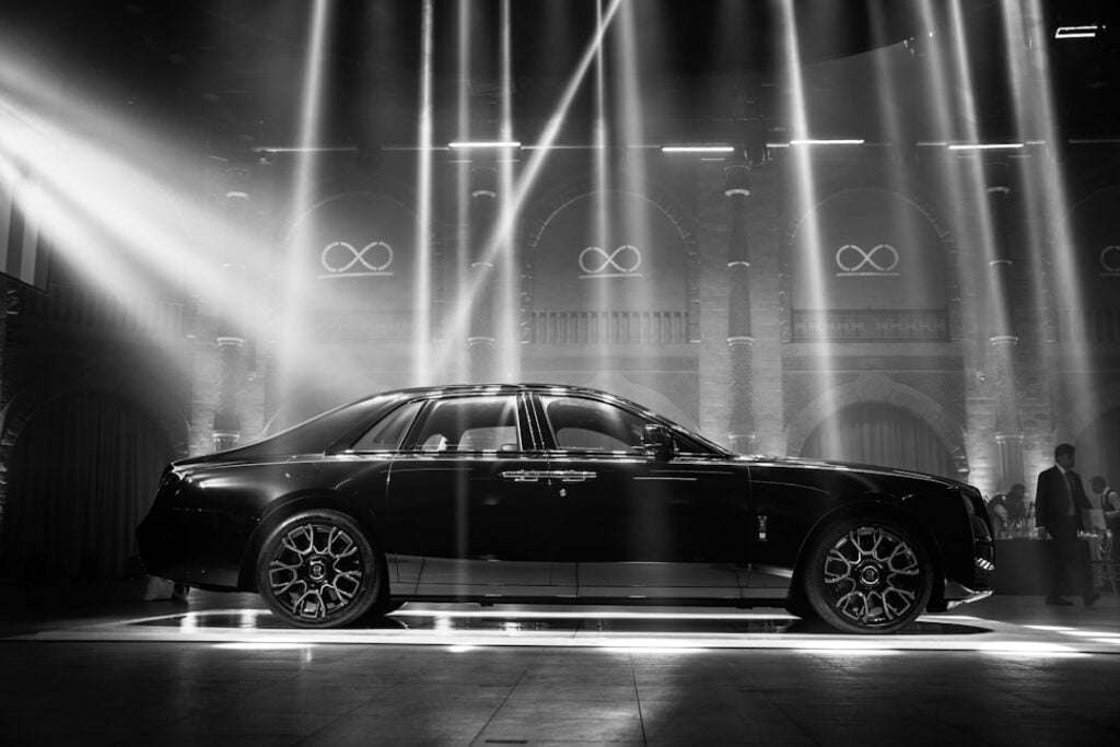 Fotoshooting für Autos: Tipps und Tricks für beeindruckende Bilder 14 Fotograf Hannover Eine schwarze Luxuslimousine wird in einem Innenbereich unter mehreren hellen Scheinwerfern ausgestellt. Die Szene ist dramatisch, mit Lichtstrahlen, die durch die leicht neblige Luft schneiden und das schlanke und elegante Erscheinungsbild des Fahrzeugs betonen. Der Hintergrund zeigt gewölbte Fenster und beleuchtete Symbole, die an die sorgfältige Einrichtung eines Autofotografen erinnern. Fotograf in der Nähe.
