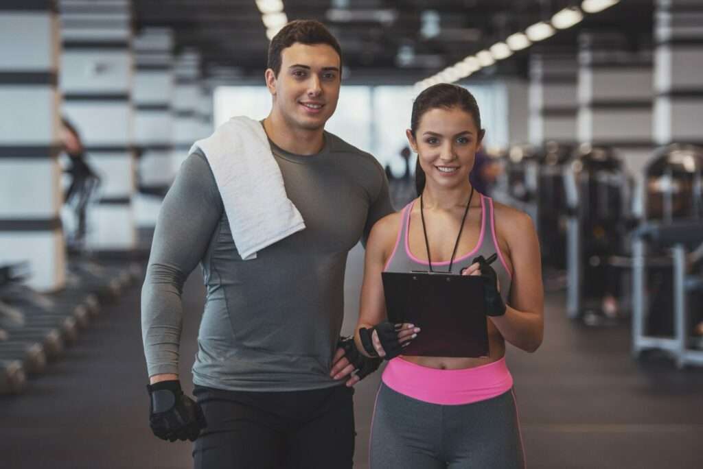 Welche Bilder darf ich gewerblich nutzen? 9 Fotograf Hannover Zwei Personen in Sportkleidung stehen in einem Fitnessstudio. Der Mann links hat ein Handtuch über der Schulter und die Frau rechts hält ein Klemmbrett. Beide lächeln. Im Hintergrund sind Trainingsgeräte zu sehen. Fotograf in der Nähe.