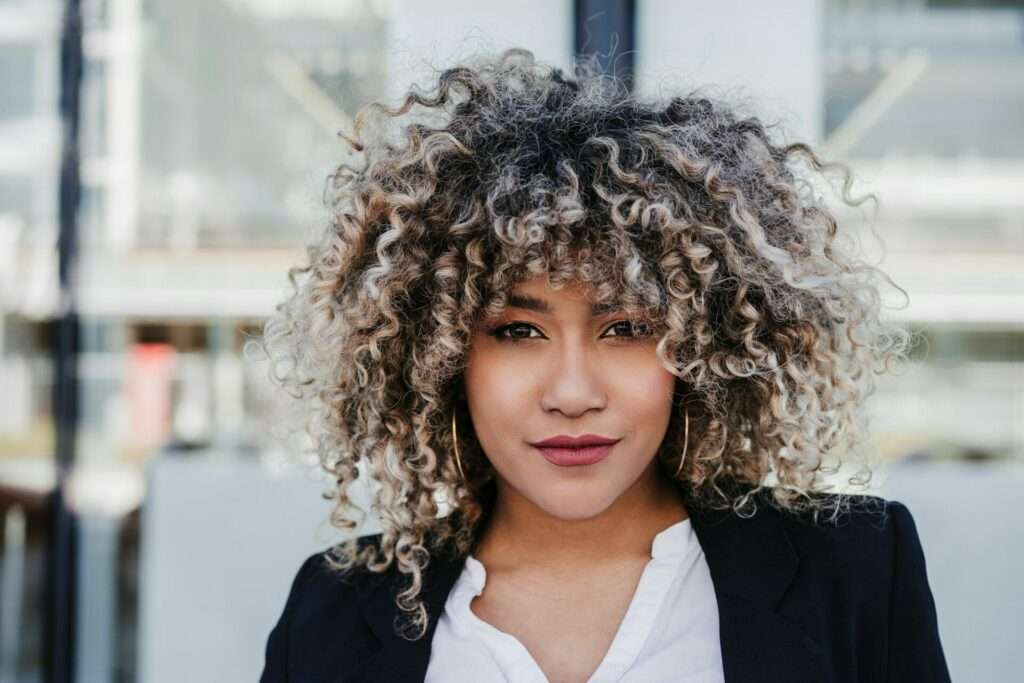 Professionelle Fotografie für Ihr Business: Businessfotos, die überzeugen 15 Fotograf Hannover Eine Frau mit lockigem Haar steht im Freien und blickt in die Kamera, um ein professionelles Geschäftsporträt aufzunehmen. Sie trägt einen schwarzen Blazer über einem weißen Oberteil und hat einen neutralen Gesichtsausdruck. Der Hintergrund ist verschwommen und zeigt ein Gebäude mit großen Fenstern. Fotograf in der Nähe.