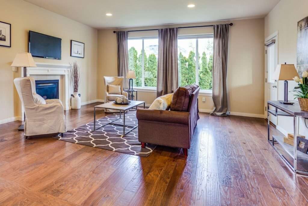 Fotograf für Immobilien gesucht: Finde den perfekten Profi! 1 Fotograf Hannover Ein helles Wohnzimmer mit einem braunen Sofa, zwei Sesseln und einem Couchtisch auf einem geometrischen Teppich ist in atemberaubenden Immobilienfotos festgehalten. Große Fenster mit Vorhängen bieten Ausblicke ins Grüne. Die Hartholzböden und der Eckkamin verleihen dem gemütlichen Raum unter einem an der Wand montierten Fernseher Wärme. Fotograf in der Nähe.