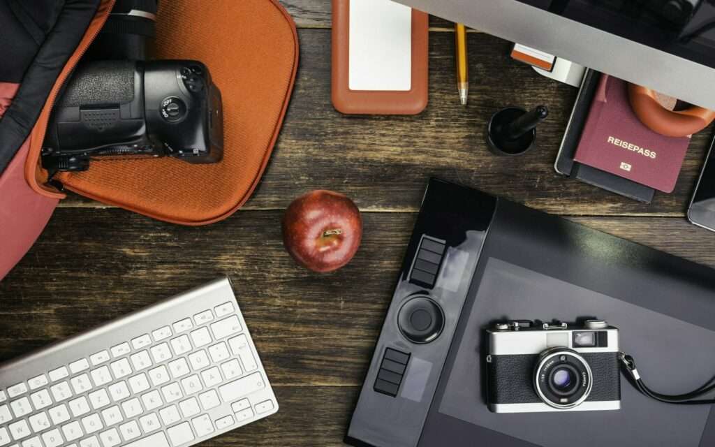 Ist Bildbearbeitung notwendig? 1 Fotograf Hannover Auf einem Holzschreibtisch stehen eine Kamera, ein digitales Tablet, eine Tastatur und eine externe Festplatte für die nahtlose Bildbearbeitung. Daneben liegen ein Apfel, ein Stift, ein Bleistift, ein roter Reisepass, ein Computermonitor und eine orange Laptophülle mit einer weiteren teilweise sichtbaren Kamera. Fotograf in der Nähe.